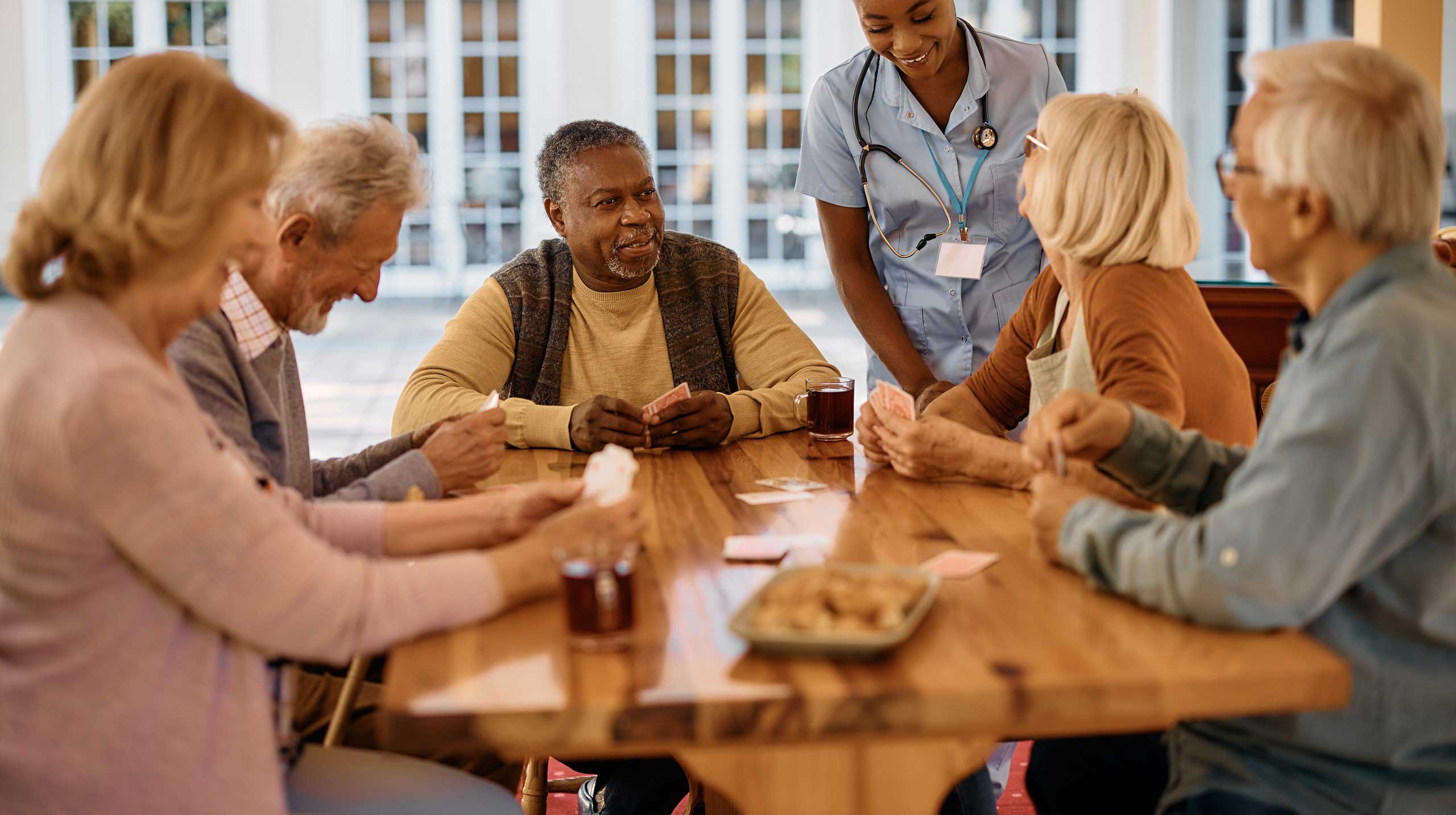 A group of seniors sit around a wooden table playing cards, with drinks and snacks, while a nurse or caregiver stands nearby smiling and talking with them, reflecting the warmth of quality senior care.