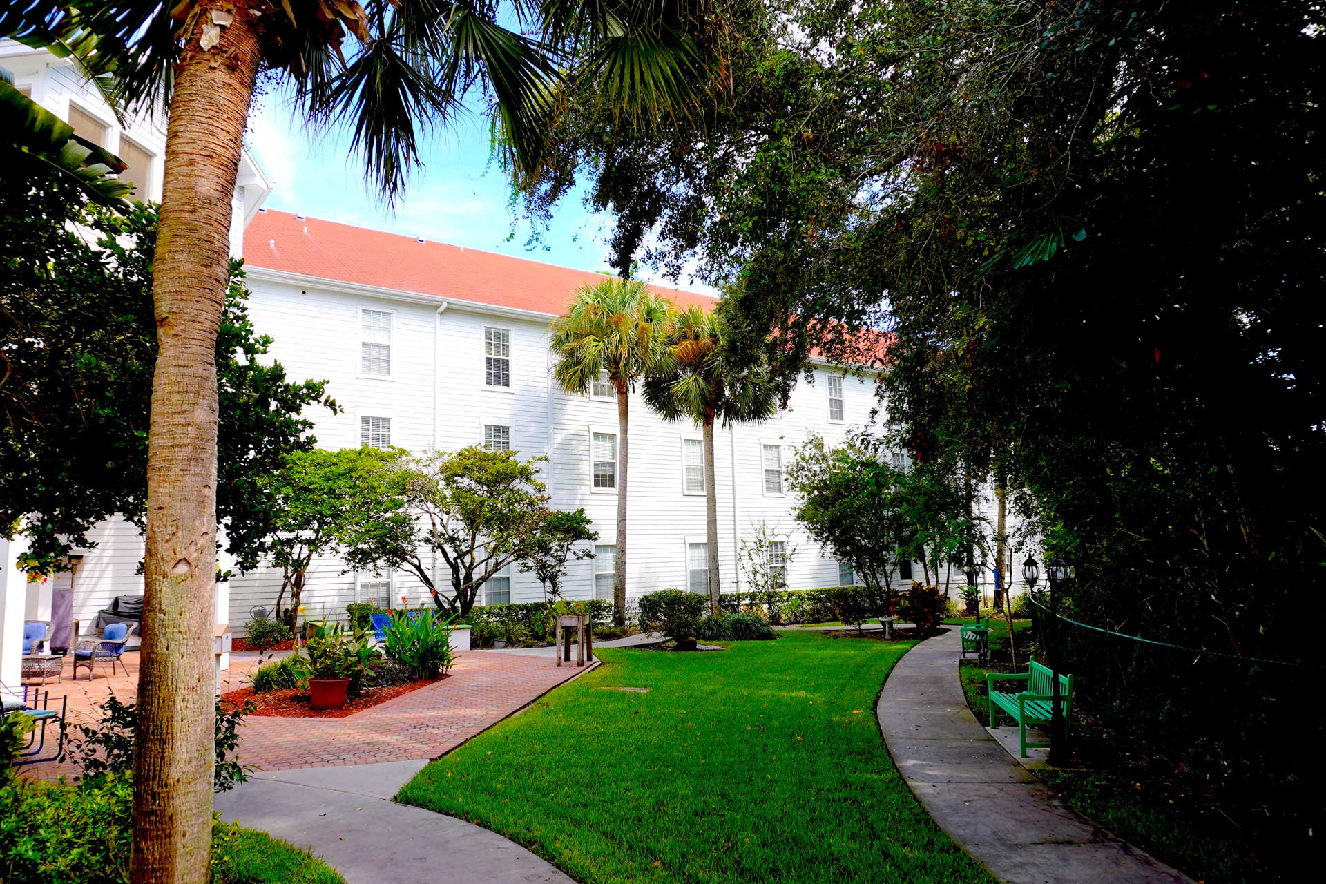 A white building with a red roof is surrounded by palm trees, green lawns, and a winding sidewalk. Benches and landscaped gardens line the shaded path, creating a tranquil setting ideal for senior care.