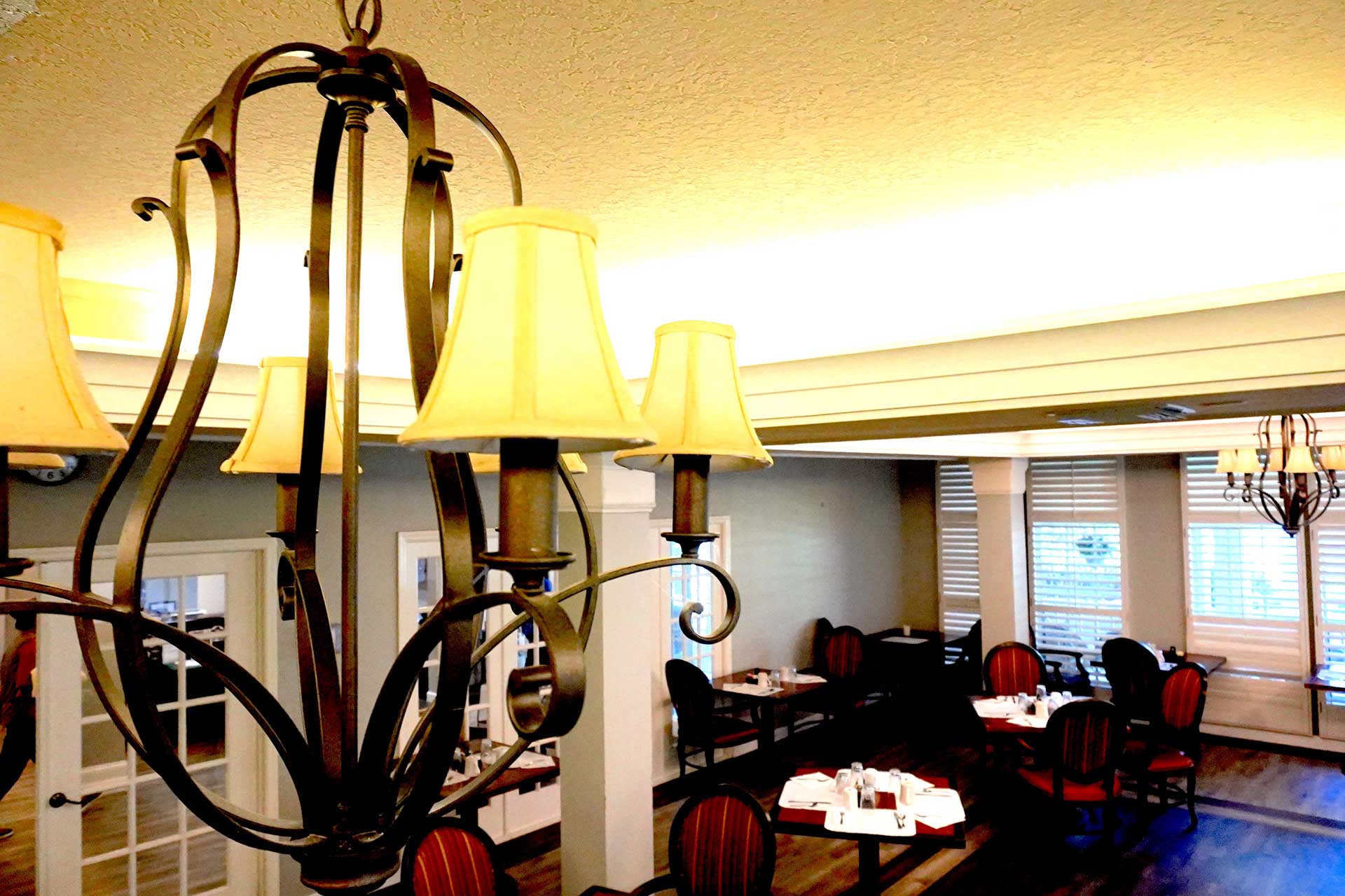 A warmly lit restaurant dining area with wooden tables and chairs, set for service. A decorative chandelier hangs in the foreground, and large windows let in natural light, creating an inviting atmosphere ideal for senior care gatherings.