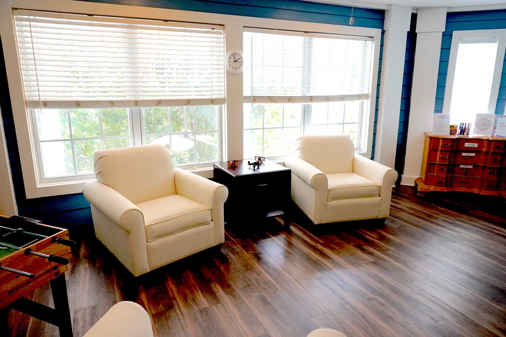 Two cream armchairs face each other beside a dark table in a bright room with large windows, wooden floors, a foosball table, and a wooden dresser. Ideal for senior care settings, sunlight streams through the white blinds, creating an inviting and comfortable atmosphere.