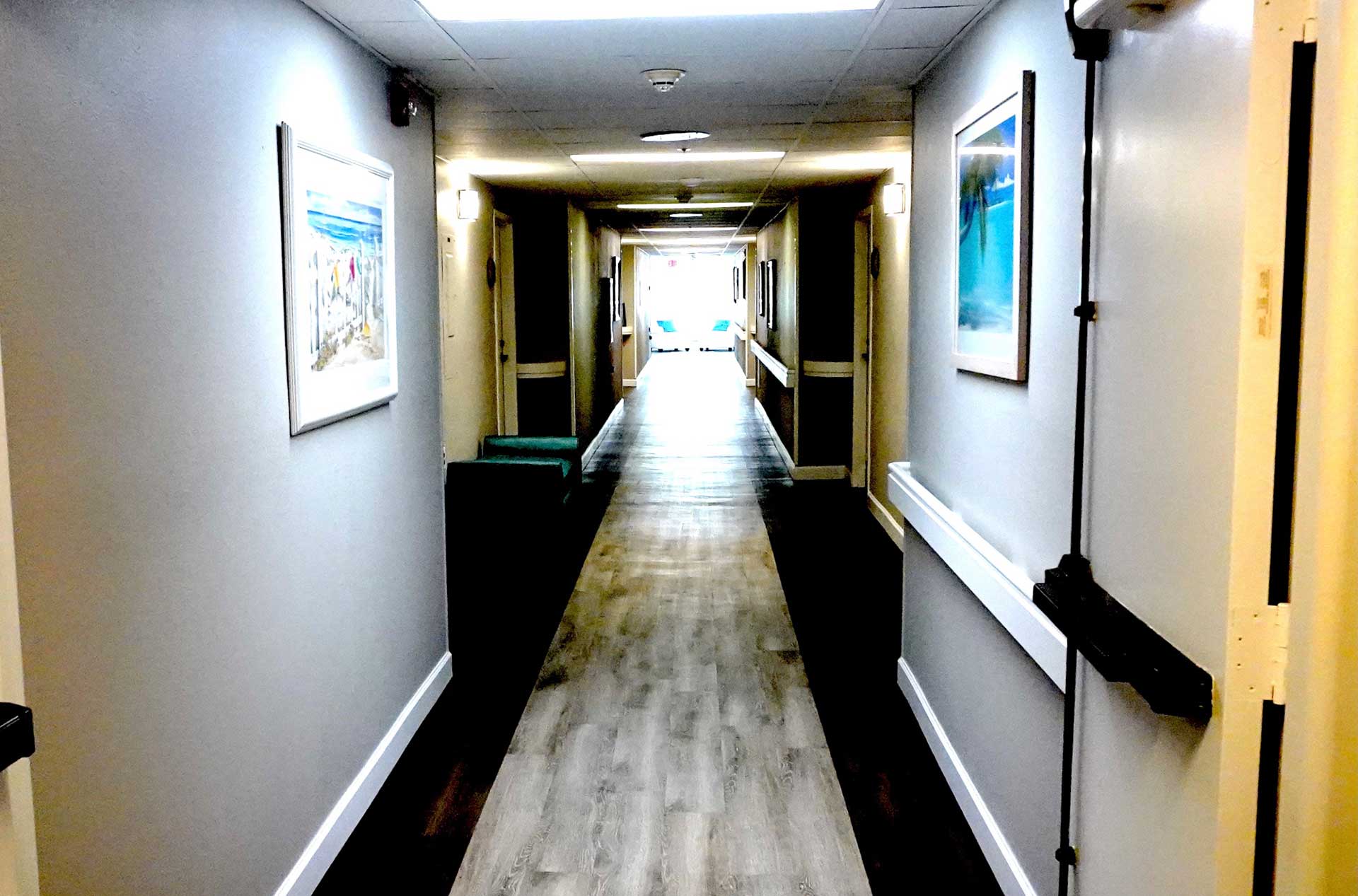A brightly lit hallway with gray walls, framed artwork, wooden floors, and white doors on each side, leading to a sunlit area at the end—designed to create a welcoming atmosphere for senior care.