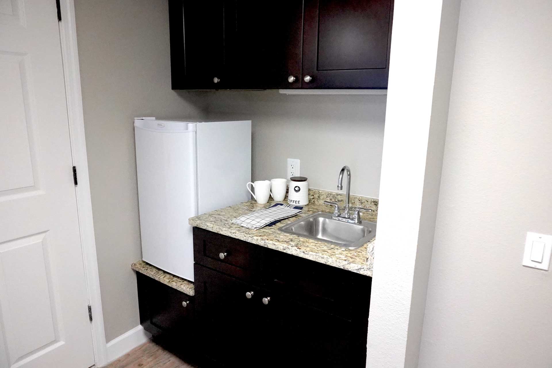 A small kitchenette designed for senior care features dark cabinets, a mini fridge, granite countertop, sink, two white mugs, a coffee container, and a dish towel. The walls are light-colored and there is a white door to the left.