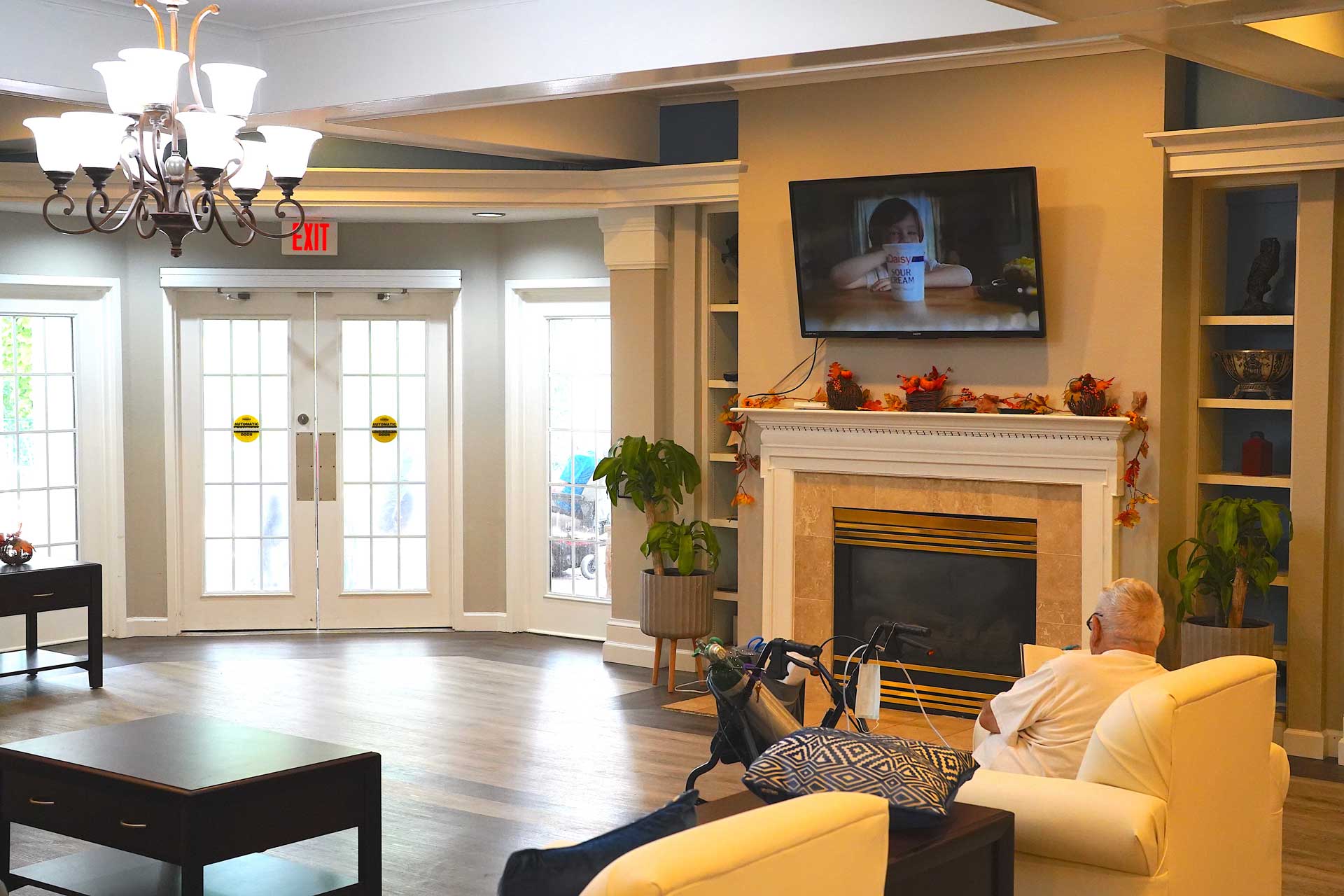 A man sits in a bright, cozy living room, watching TV above a fireplace decorated with autumn leaves. The space features chairs, a table, and large windows letting in natural light, creating an inviting atmosphere ideal for senior care.