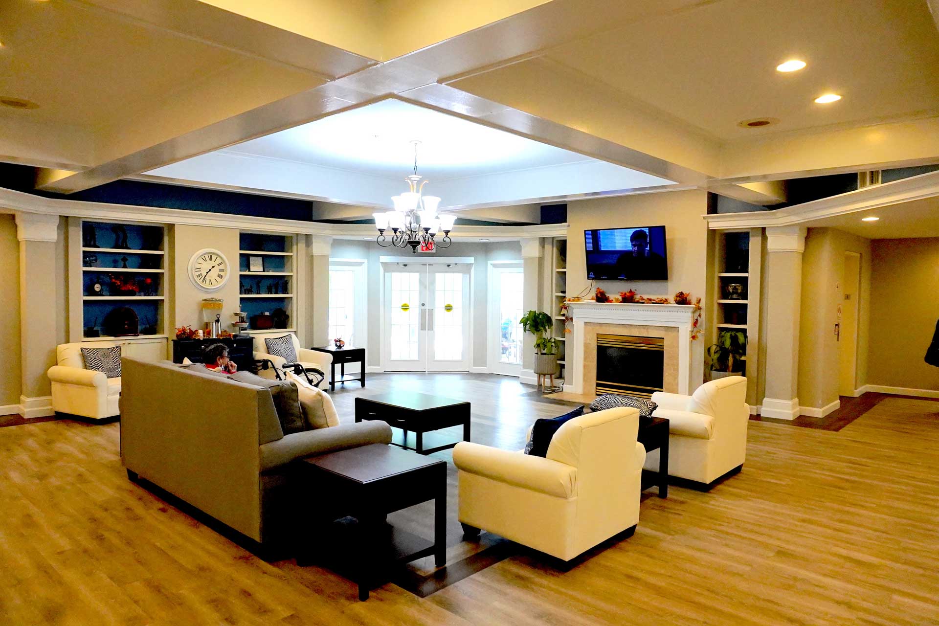 A spacious, well-lit living room with beige sofas, a fireplace, built-in shelves, a chandelier, wooden floors, and large windows letting in natural light—an inviting setting perfectly suited for comfortable senior care.