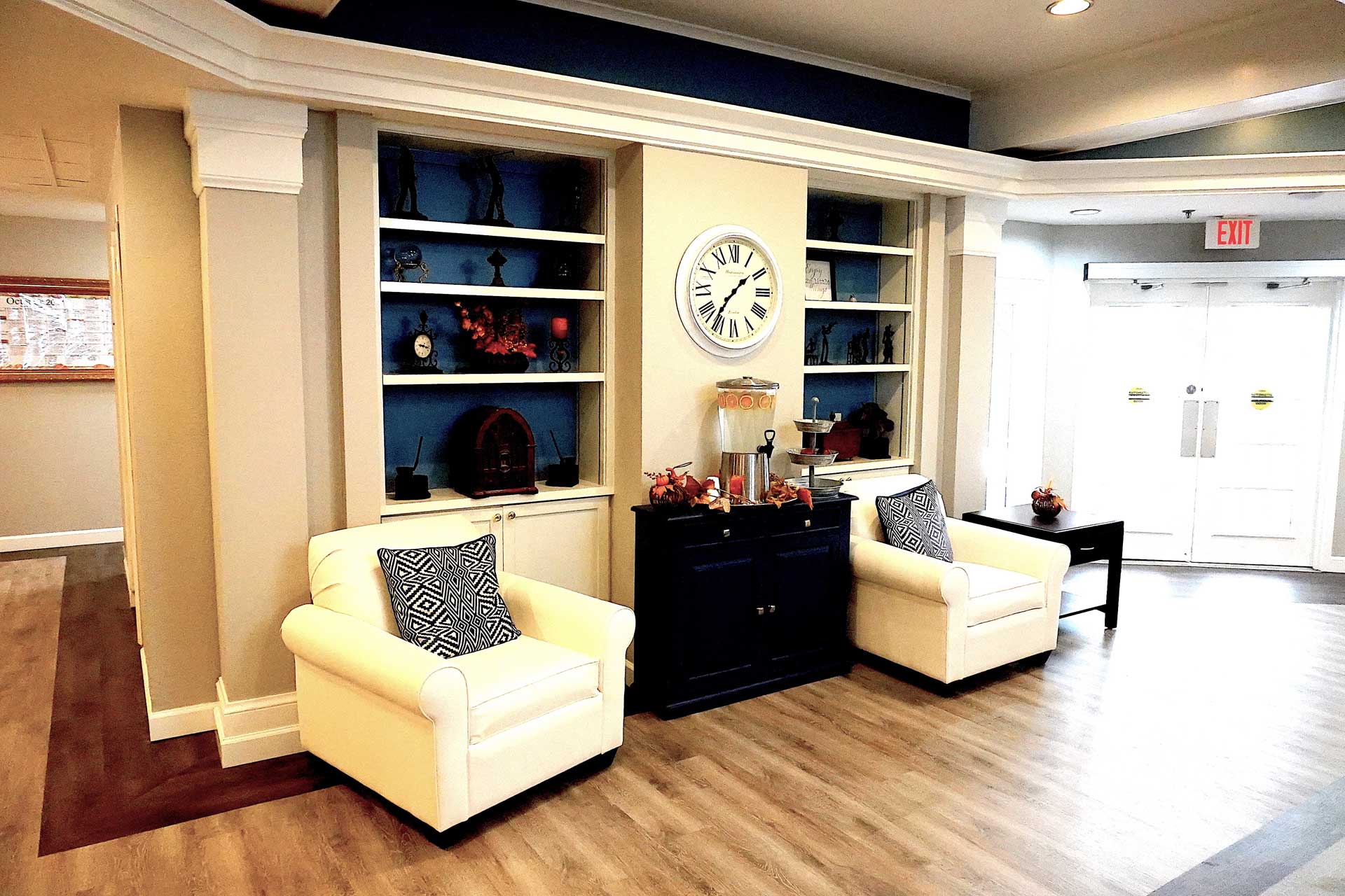 A bright lobby area designed for senior care features two white armchairs, a black chest, bookshelves, a wall clock, and decorative items. Glass doors let in natural light from the right.