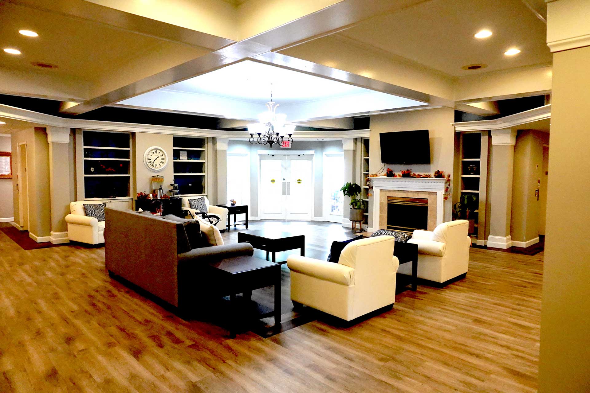 A spacious, well-lit living room designed with senior care in mind, featuring wood flooring, cream armchairs, a gray sofa, fireplace, TV, bookshelves, chandelier, and double glass doors at the back.