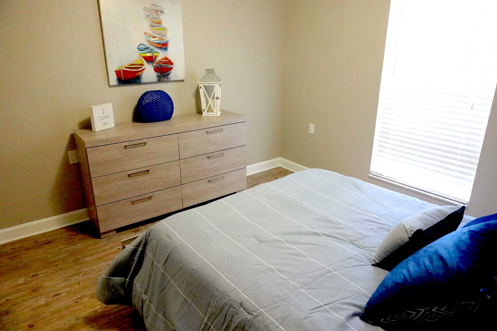 A neatly made bed with gray bedding and blue pillows sits beside a window. A wooden dresser holds a blue vase, a white lantern, and framed art. Reflecting thoughtful senior care, a colorful boat painting hangs above the dresser.