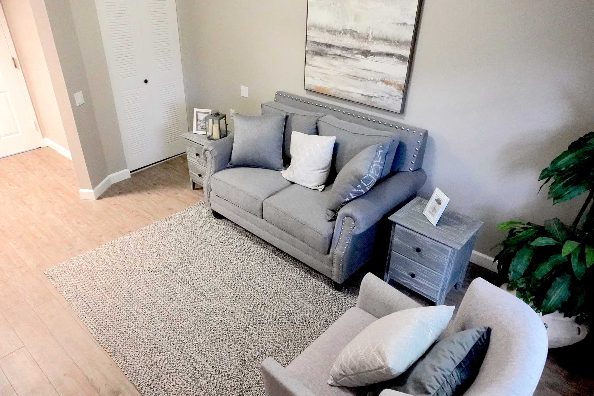 Gray loveseat and armchair with blue-gray pillows, matching side tables, a textured area rug, abstract wall art, and a potted plant create a comfortable and inviting atmosphere in this modern living room with wooden floors—an ideal setting for senior care.