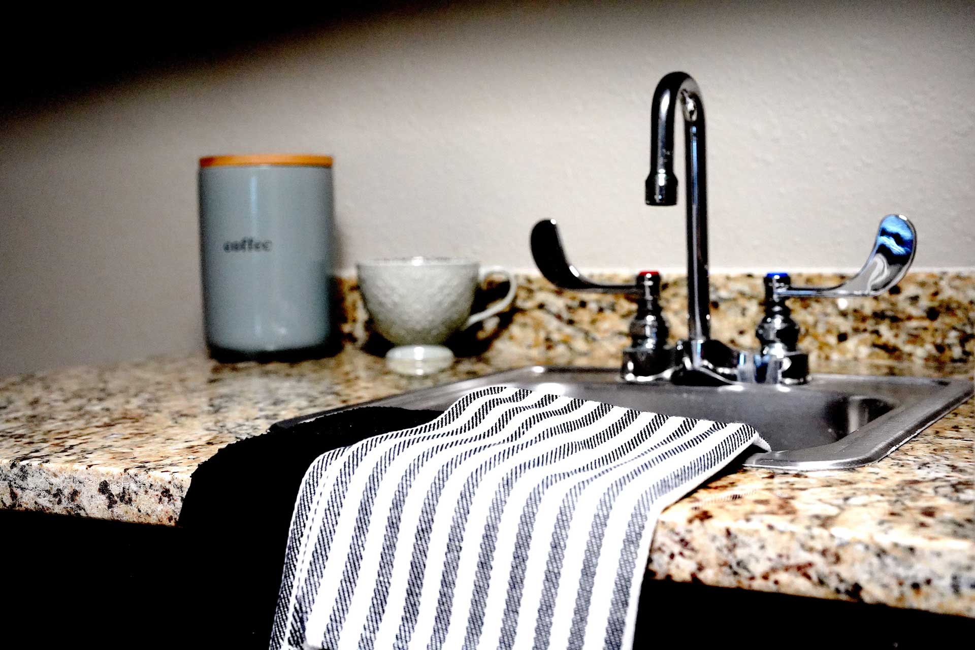 A striped dish towel is draped over a granite countertop next to a kitchen sink, with a coffee canister and a white teacup placed in the background—creating an inviting space that’s ideal for senior care meal preparation.