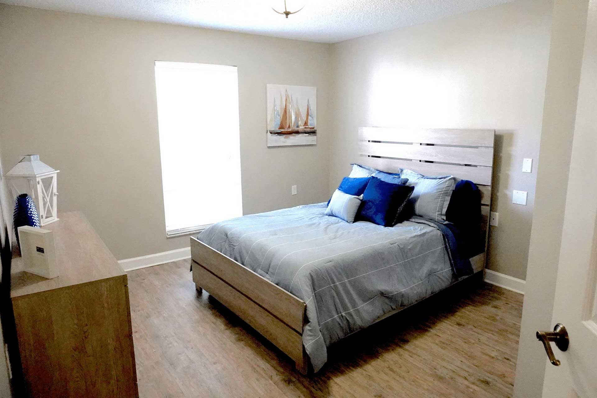 A simple bedroom designed for senior care features a wooden bed, blue and gray bedding, a dresser, a window with blinds, and a sailboat painting on the beige wall. The floor is light wood, and the room is well-lit.