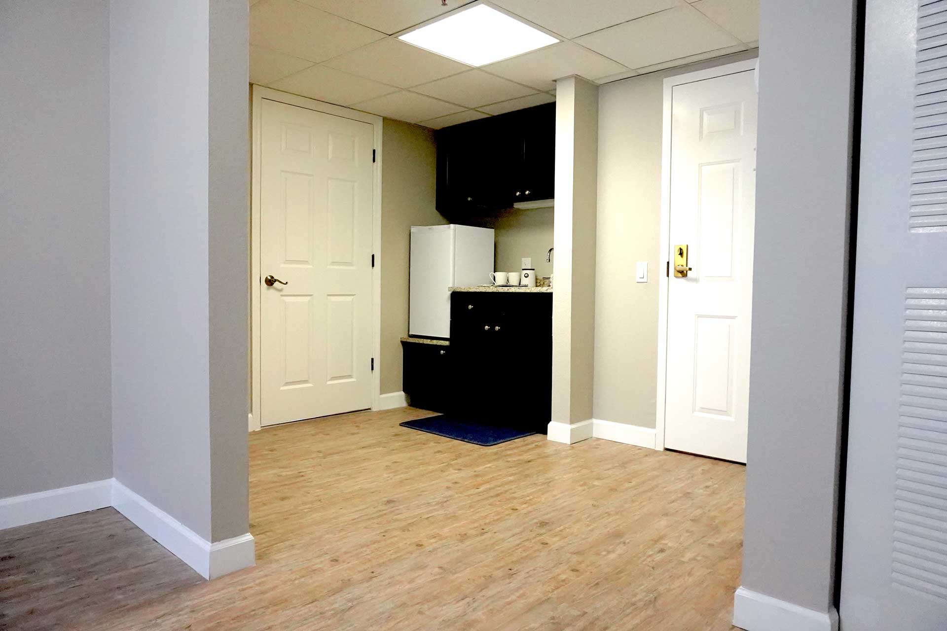 Small kitchenette with a white mini fridge, dark cabinets, sink, and blue mat. Two closed white doors and light wood flooring are visible in a brightly lit, neutral-colored room, ideal for senior care settings.