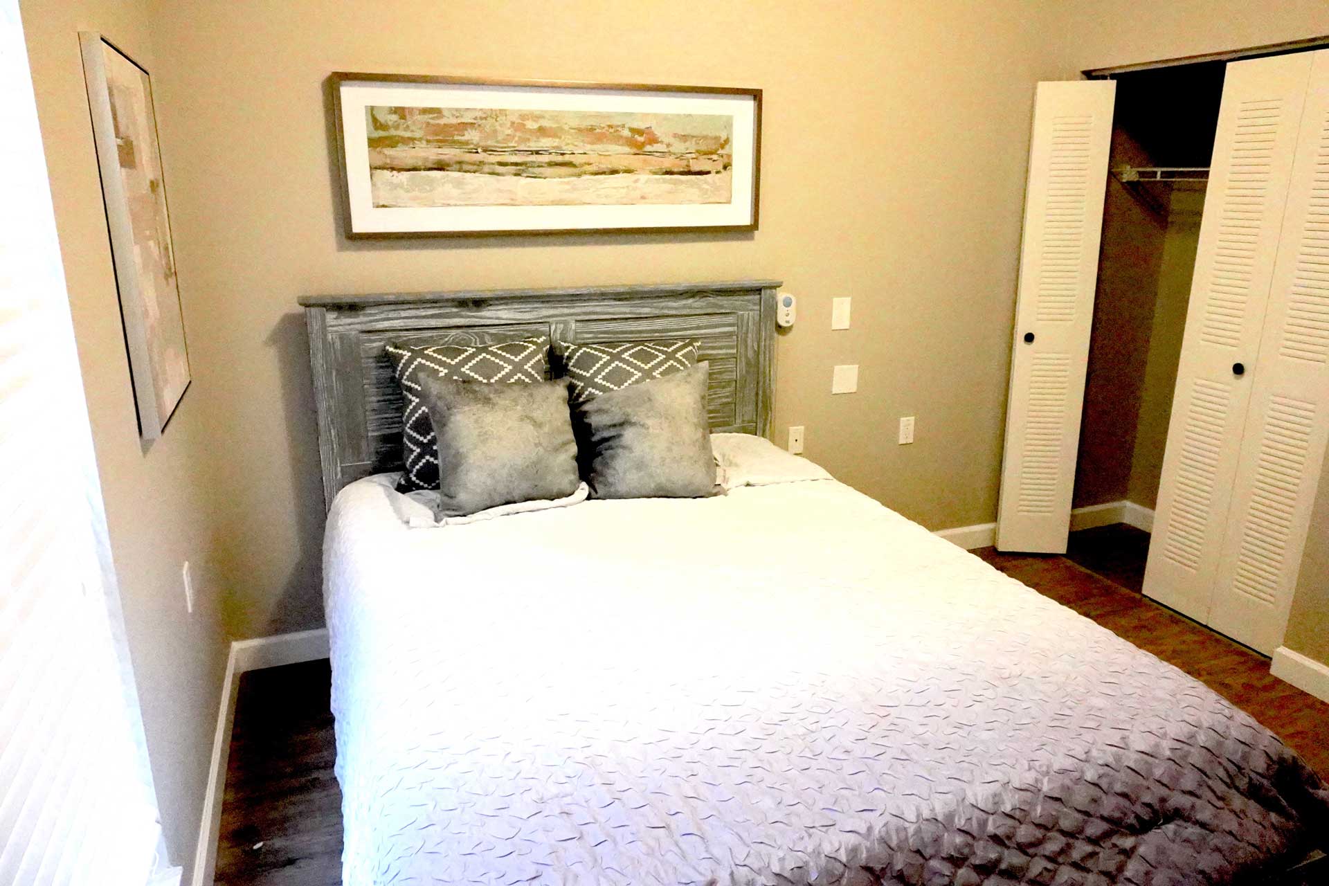 A neatly made bed with gray pillows sits against a beige wall under a framed picture, creating a welcoming atmosphere suitable for senior care. The room features wooden floors, an open closet with white doors, and natural light streaming in from the left.