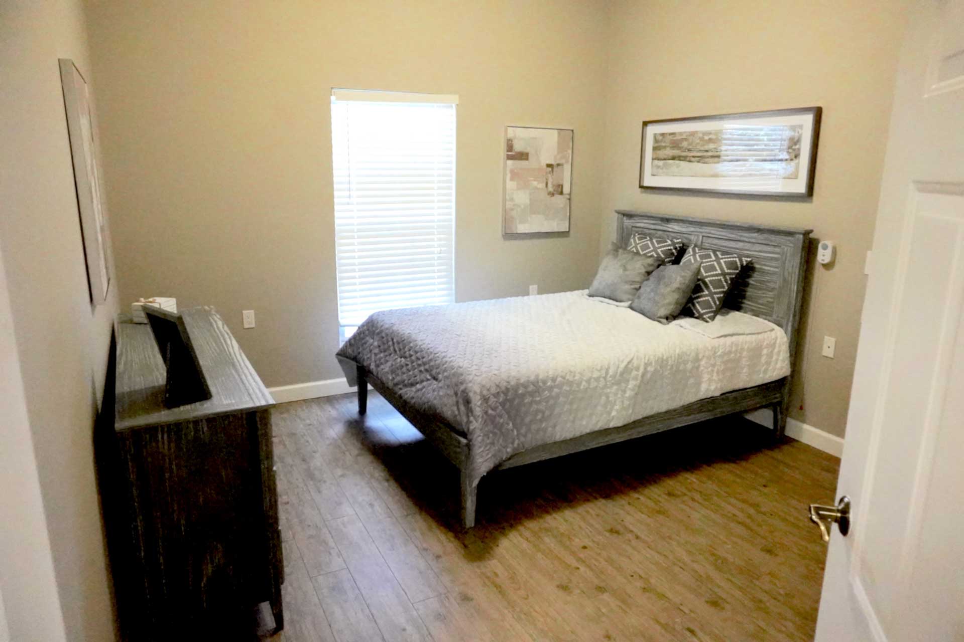 A small bedroom designed for senior care features a wooden bed with gray bedding and three pillows, a dresser topped with a TV, wooden flooring, a window with blinds, and framed art decorating the beige walls.