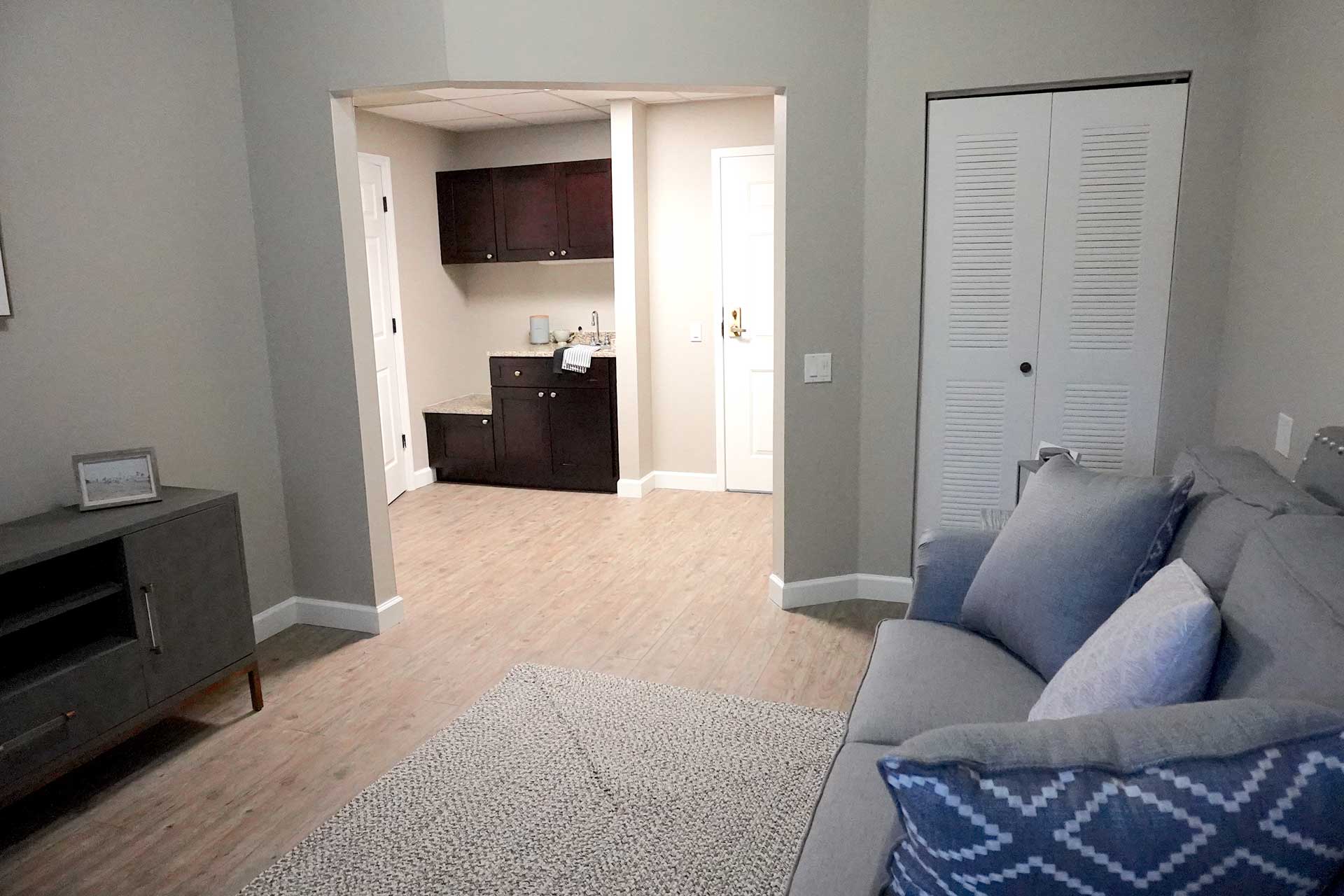 A modern living room designed for senior care features a gray sofa, blue and white pillows, light wood flooring, and an open doorway leading to a kitchenette with dark cabinets. A closed closet door is on the right.