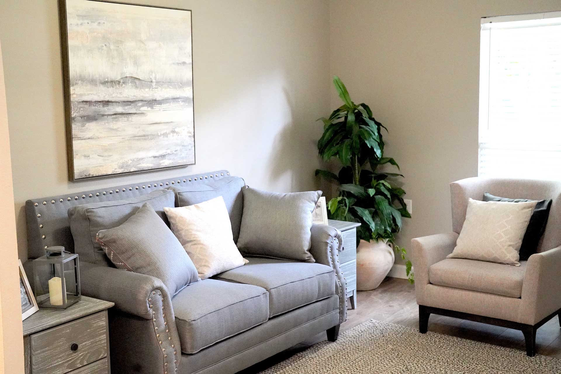 A cozy living room designed for senior care features a gray loveseat, an armchair, a side table, a large potted plant, wall art, and a window letting in natural light. Neutral colors and soft textures create a calm atmosphere.