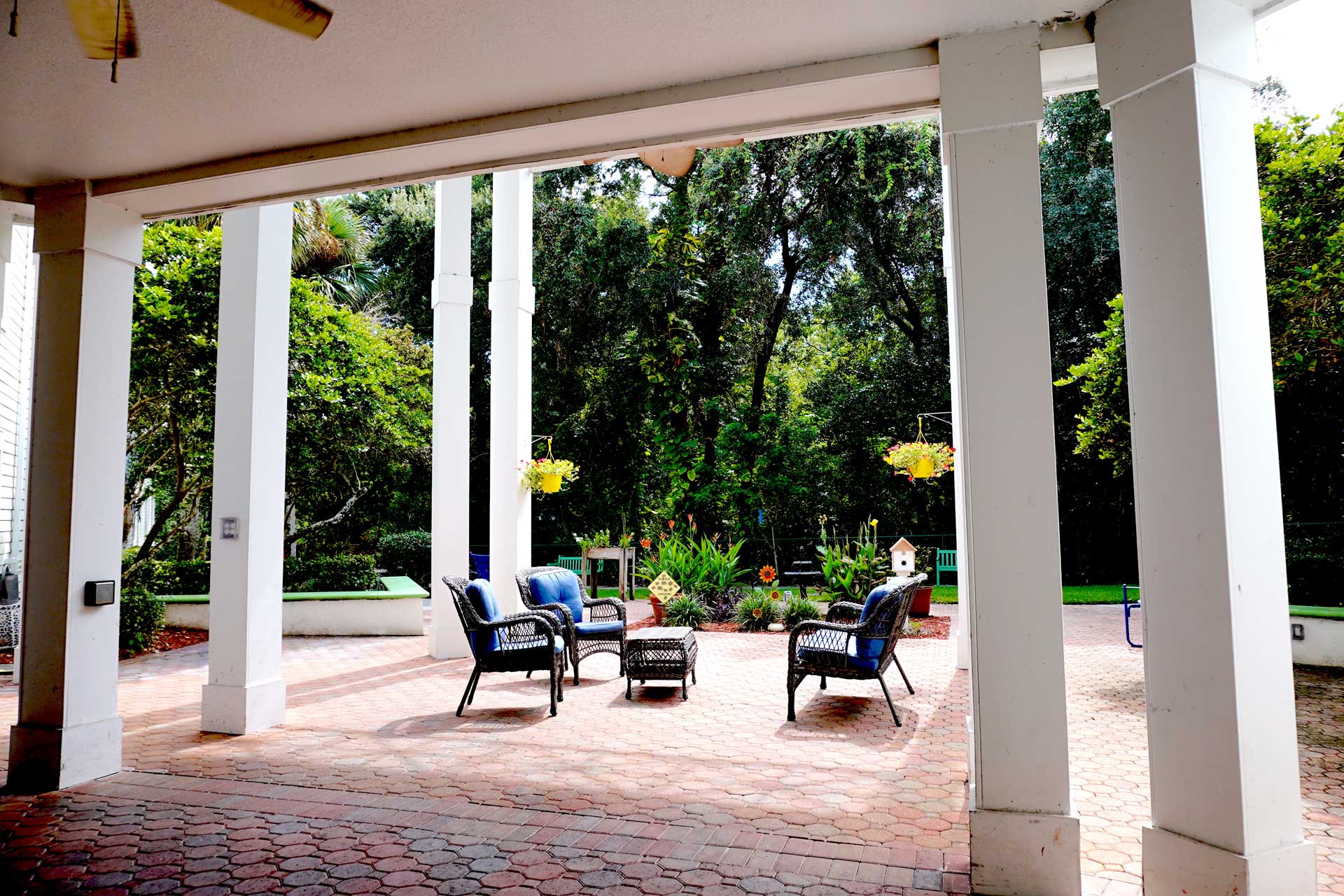 Outdoor patio with four cushioned chairs and a table arranged on brick flooring, surrounded by white columns, greenery, and hanging flower baskets in a backyard setting—ideal for comfortable senior care gatherings or relaxation.