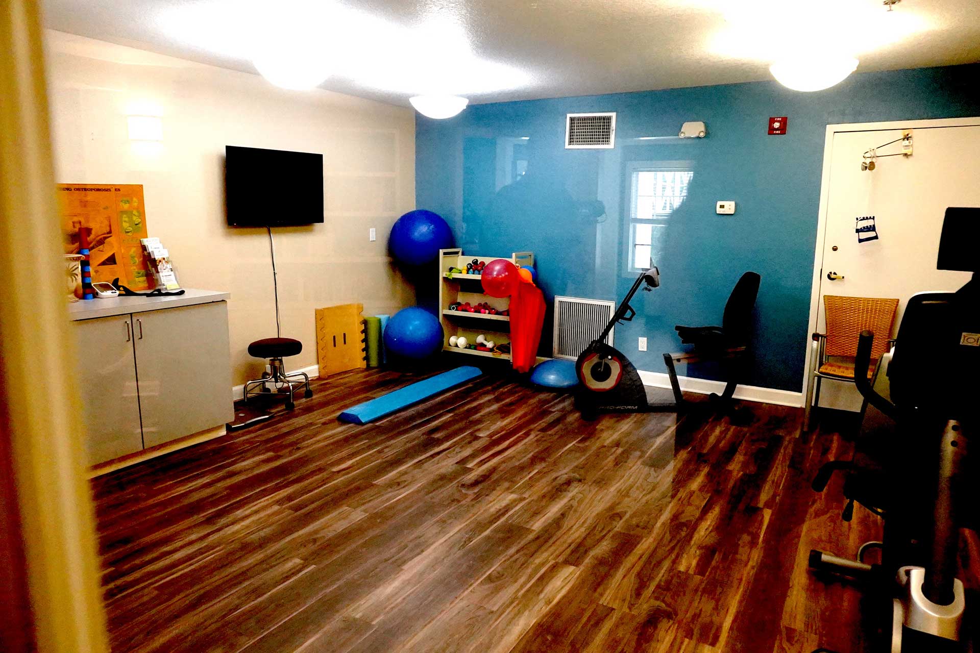 Small gym with wood floors, an exercise bike, yoga mat, exercise balls, a TV, shelving with fitness equipment, and a blue accent wall—thoughtfully designed to support senior care and wellness activities.