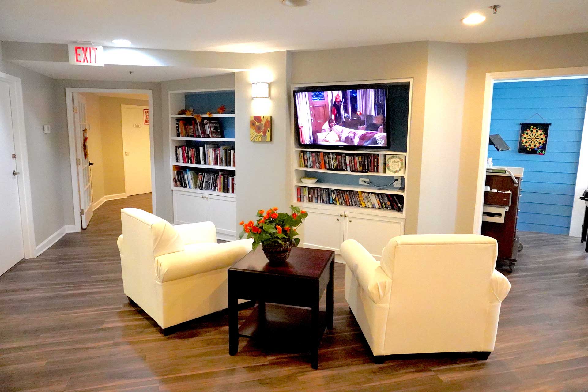Two white armchairs face a TV in a cozy room with bookshelves, a table with flowers, and wood flooring. A dartboard and piano are visible in adjacent rooms, creating inviting spaces perfect for senior care. Bright lighting gives the space a welcoming feel.