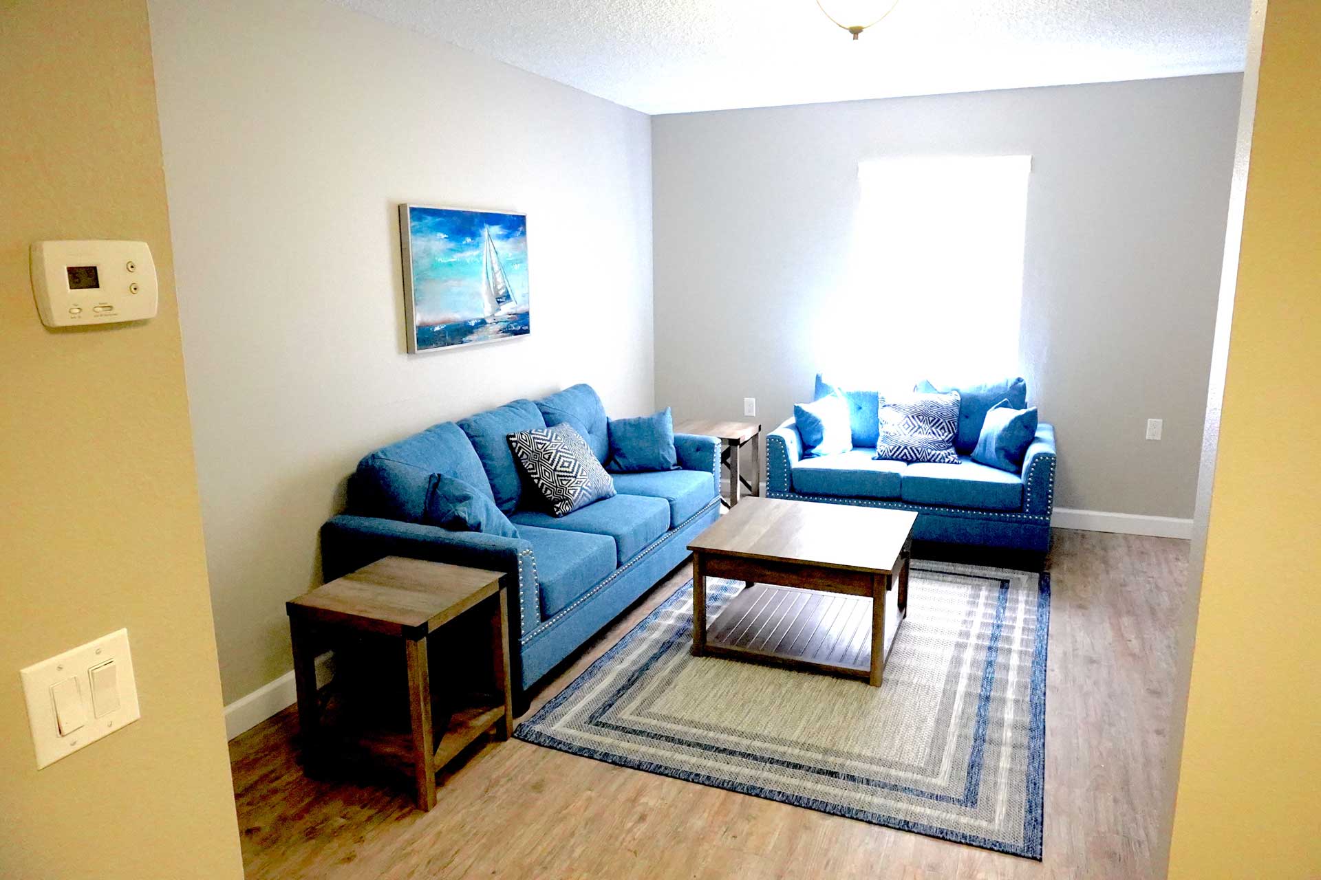 A small living room designed for senior care features two blue couches, a wooden coffee table, an end table, a rug, and a window letting in light. A boat painting hangs on the wall above the couch.