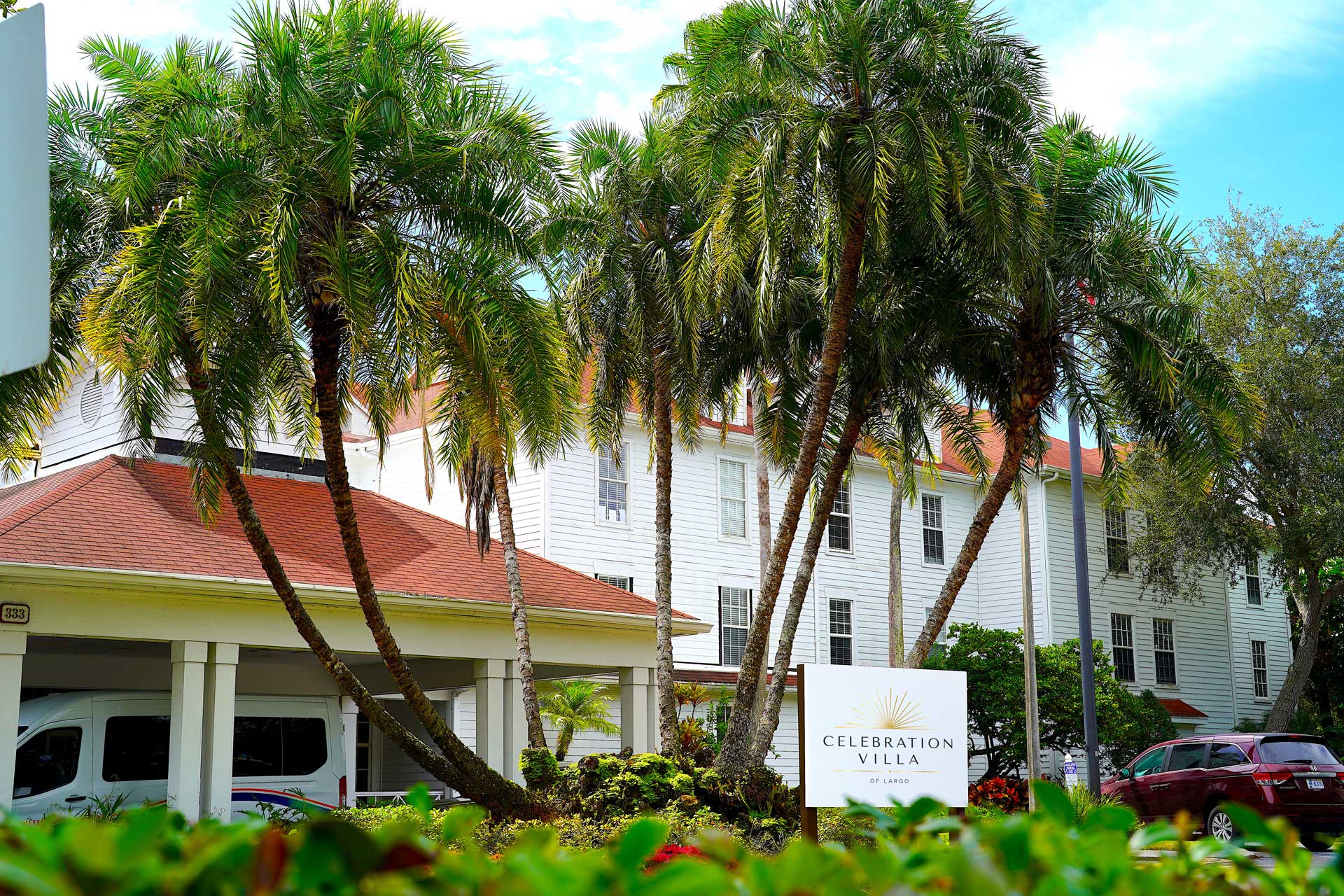 A white building with a red roof stands behind tall palm trees and a sign that reads “Celebration Villa.” Shrubs and greenery are in the foreground, and a van is partially visible to the left, highlighting the welcoming atmosphere of this senior care community.