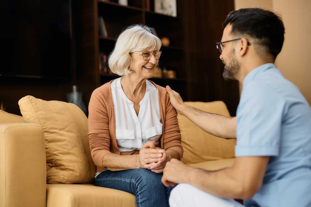 assisted_living_585799768 A woman sits on a couch smiling as a younger man with glasses gently touches her shoulder. Both appear happy and engaged in conversation in a cozy living room, reflecting the warmth and companionship found in quality senior care.