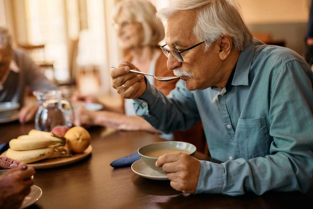 assisted_living_experience_delight_521989096 A man with glasses eats soup at a table, while a woman sits beside him. There is a plate of bananas and apples in the background, creating a warm atmosphere that reflects senior care.