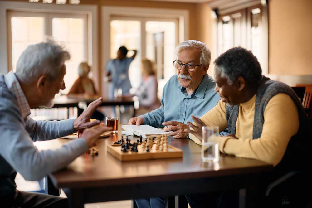 Three men sit at a table, playing chess and talking. Two glasses of water and a cup of tea are on the table. More people are visible in the background of a bright, cozy room, creating an inviting atmosphere ideal for senior care activities.
