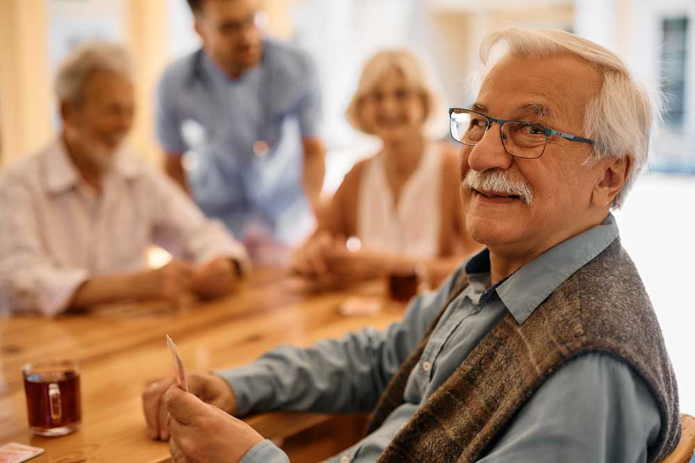 A man with glasses smiles at the camera while holding playing cards at a table with three other seniors. They appear to be enjoying a game together in a bright, cheerful room, reflecting the positive atmosphere of senior care.