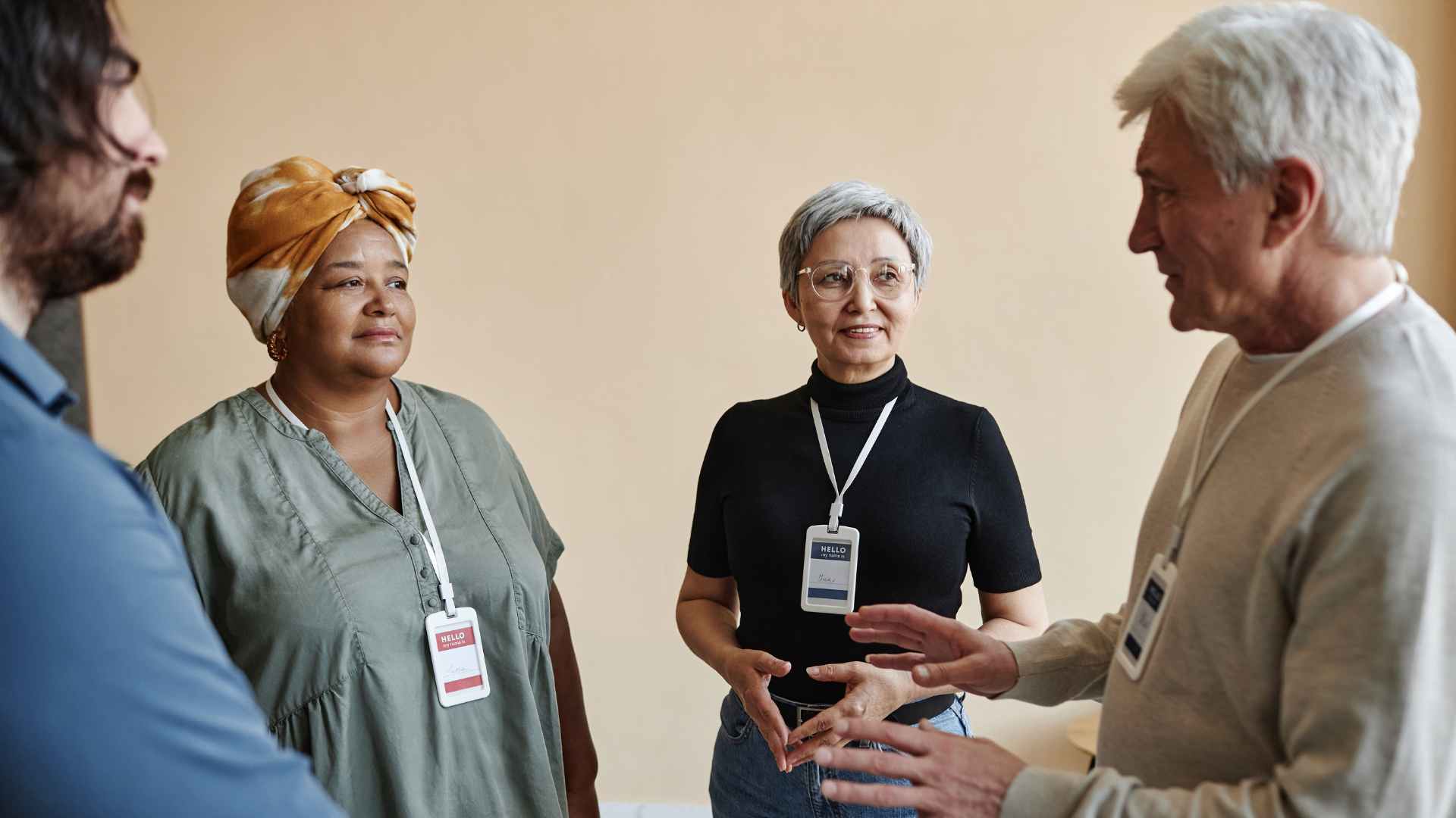 Four adults wearing name badges stand in a circle having a discussion about senior care. One man gestures while speaking as the other three people listen attentively. The background is neutral and simple.