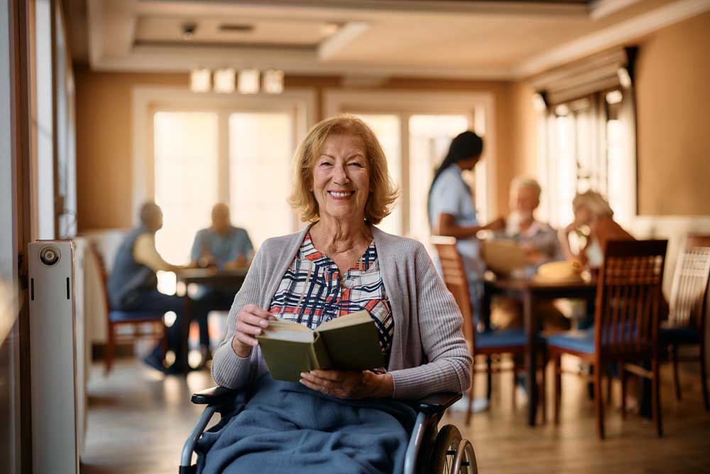 Smiling woman in a wheelchair reads a book in a bright room, with other seniors and caregivers interacting at tables in the background, illustrating the warmth of senior care.